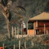 Homem parado ao lado de uma cabana de madeira com vegetação tropical ao fundo. Imagem ilustra artigo do Barrazine (Os melhores lugares para estar em contato com a natureza na Barra da Tijuca).