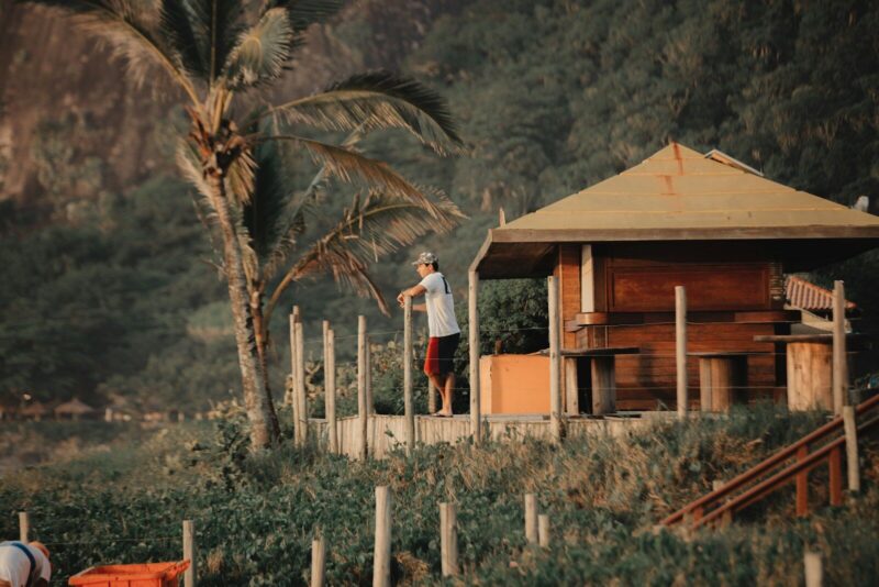 Homem parado ao lado de uma cabana de madeira com vegetação tropical ao fundo. Imagem ilustra artigo do Barrazine (Os melhores lugares para estar em contato com a natureza na Barra da Tijuca).
