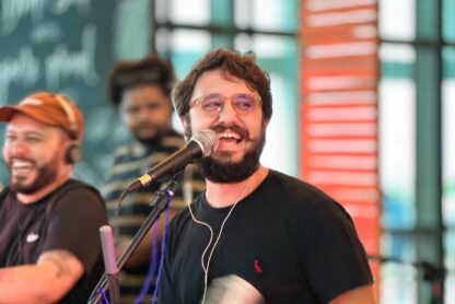 Um homem sorridente e barbudo cantando ao microfone, com outro músico sorrindo ao fundo, em um local com janelas verdes ao fundo.